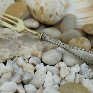 May include: A silver-colored fork with a decorative handle, resting on a bed of white and beige stones.