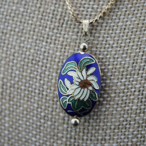May include: A gold chain necklace with a blue and white enamel pendant featuring a floral design. The pendant is oval shaped and has a gold trim.