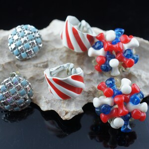 May include: A set of five earrings featuring a variety of styles. Two pairs of earrings are made of white and red striped metal. One pair is made of blue and white squares. The other two pairs are made of red, white, and blue beads.