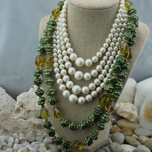 May include: A multi-strand necklace set on a display stand. The necklace features layers of white faux pearls and green beaded strands with gold accents. The beads are olive green and the necklace has a vintage aesthetic.