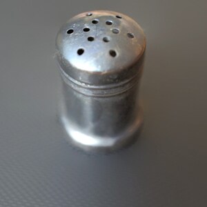 Może przedstawiać: Srebrna solniczka z okrągłym wierzchołkiem i szeregiem otworów do dozowania soli.