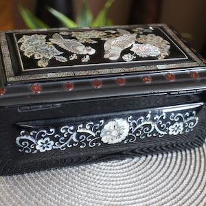Może przedstawiać: Czarne drewniane pudełko z ozdobnym wzorem kwiatów i ptaków inkrustowanych perłową skorupą. Pudełko ma zawiasowe wieczko i jest zamknięte.