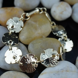 May include: Gold-toned bracelet featuring a chain with multiple round, textured, sand dollar-shaped charms. The bracelet is photographed on a bed of smooth, light brown stones.