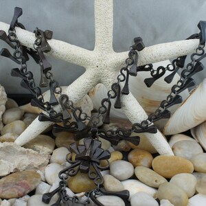 May include: A black metal necklace with a large pendant made of multiple metal pieces. The necklace is displayed on a white starfish and surrounded by white and brown stones.