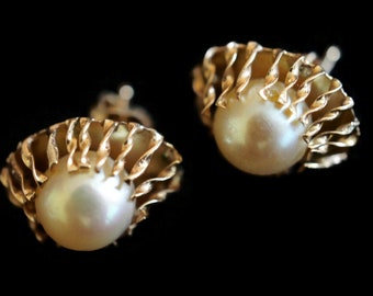 May include: A pair of gold-toned earrings, each featuring a large, lustrous pearl. The pearls are cradled within a decorative, swirling gold setting, creating a seashell-like appearance. The background is a solid black.