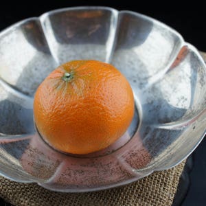 May include: A silver, flower-shaped bowl with a single orange in the center.