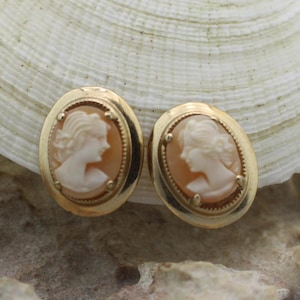 May include: A pair of gold tone cameo earrings with a white cameo of a woman's profile. The earrings are oval shaped and have a gold tone border.