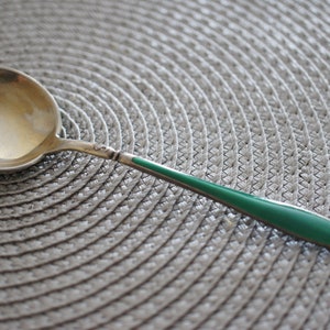 May include: A silver spoon with a green enamel handle. The spoon has a rounded bowl and a decorative design on the handle.