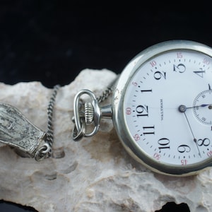 Op de afbeelding: Een zilveren zakhorloge met een witte wijzerplaat en zwarte Romeinse cijfers. Het horloge heeft een kleine secondewijzer-wijzerplaat en de woorden "Waltham" staan ​​gedrukt op de wijzerplaat. Het horloge is bevestigd aan een zilveren ketting met een decoratieve sluiting.