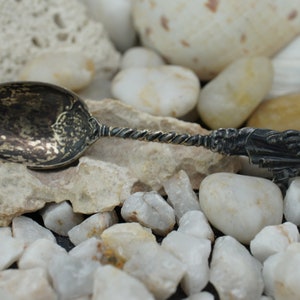 May include: An antique silver spoon with an ornate handle and a twisted stem. The spoon is resting on a bed of white and beige stones, with a seashell in the background. The spoon's bowl has a tarnished appearance.