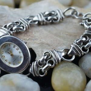 Op de afbeelding: Een zilveren horloge met een ovale wijzerplaat en een zilveren kettingarmband. De wijzerplaat van het horloge heeft Romeinse cijfers als uurmarkeringen en een zilveren secondewijzer.