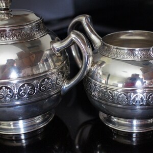 Può includere: Un set da tè in argento con una teiera e un creamer. Entrambi i pezzi hanno un design ornamentale con un bordo rialzato.
