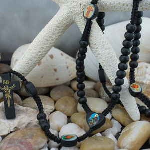 May include: Black rosary with wooden beads and a crucifix. The rosary features small, round, and heart-shaped decorative elements with religious imagery. The background includes seashells and small stones, creating a natural aesthetic.