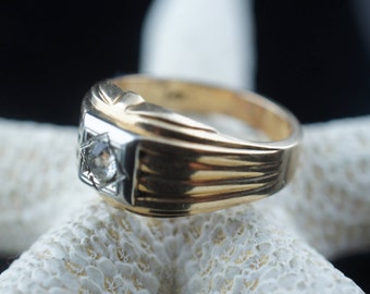 May include: A gold ring with a single diamond set in a white metal setting. The ring has a textured band with a grooved design.