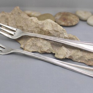 May include: Two silver forks with three tines each, resting on a piece of light brown rock. The forks have a simple, elegant design with a decorative element at the end of the handle. The background includes small stones and a seashell.