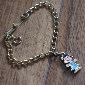 May include: A gold chain bracelet with a charm of a girl wearing a blue dress. The charm is attached to the chain with a small ring.