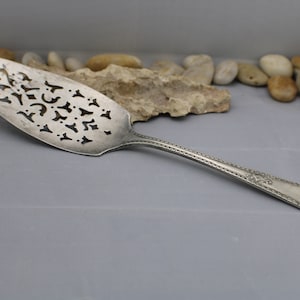 Pode incluir: Um servidor de bolo de prata com um design decorativo recortado. O servidor tem um cabo com contas e uma borda festoneada.