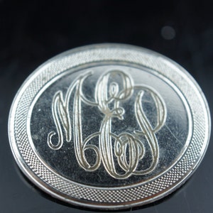 Puede incluir: Un compacto redondo plateado con un diseño monogramado. El monograma está en una fuente de escritura y dice "M.E.B."