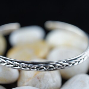 May include: A silver bangle bracelet with a braided design. The bracelet is on a white background.