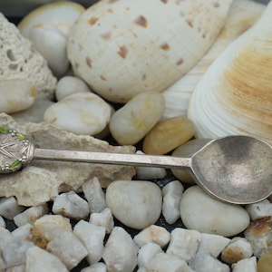 May include: A silver teaspoon with a green and white enamel star design on the handle. The spoon is resting on a bed of white and brown stones.