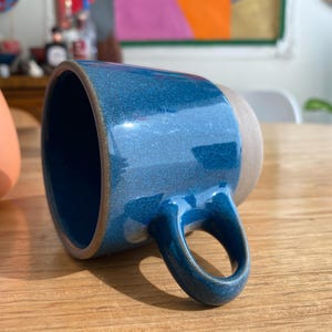 May include: A handmade ceramic mug with a glossy blue glaze and a natural, unglazed base. The mug has a rounded handle and is set on a wooden surface. Another orange ceramic object is visible in the background.