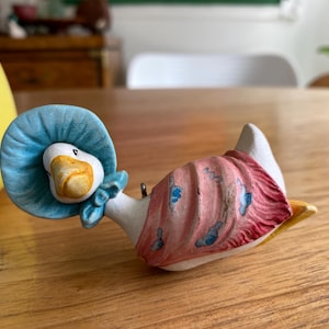 May include: A ceramic goose figurine wearing a blue bonnet and a pink shawl with blue butterfly accents. The goose has a yellow beak and feet. The figurine is on a wooden surface.