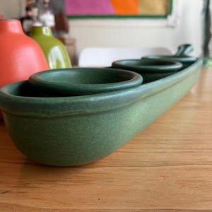 May include: A green ceramic serving dish with three round bowls set in a long, oval-shaped tray. The dish is a muted green color and sits on a wooden surface. In the background are three colorful vases.
