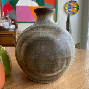 May include: A handcrafted, round ceramic vase with a wide body and a narrow neck. The vase features a textured, earthy color palette of brown, tan, and cream, with a darker brown rim. The vase is displayed on a wooden table.