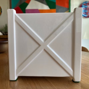 May include: White ceramic planter with a square shape and a criss-cross design on the front. The planter has green rubber feet on the bottom.