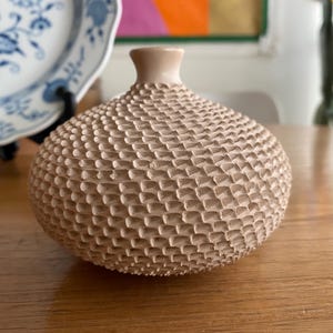 May include: A small, beige ceramic vase with a textured, patterned surface. The vase has a rounded body and a narrow neck. The vase is sitting on a wooden table. Decorative home decor.