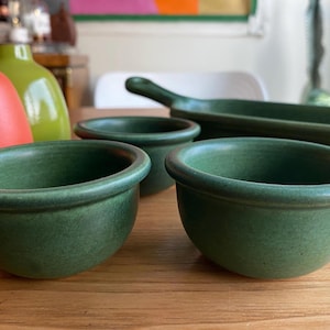 May include: A collection of green ceramic bowls and a rectangular dish on a wooden surface. The bowls have a rounded shape with a slightly raised rim. In the background, there are two colorful bottles.