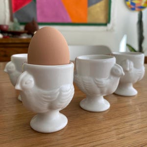 May include: A brown egg sits in a white ceramic egg cup, with three other egg cups nearby. The egg cups are shaped like chickens, with detailed feathers and a small beak. The cups are on a wooden table.