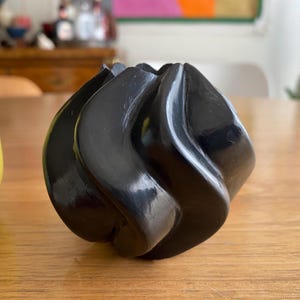 May include: A black, sculptural vase with a swirling, organic design. The vase has a glossy finish and a unique, abstract shape. The vase is sitting on a wooden surface.