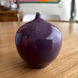 May include: A glossy, deep purple vase with a rounded body and a slender neck. The vase reflects the surrounding environment, creating a play of light and shadow. The ceramic vase sits on a wooden surface.