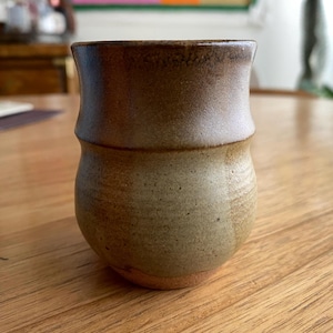 May include: A handmade ceramic tumbler with a two-tone design. The top portion is a dark brown, transitioning to a lighter tan-brown at the base. The cup has a rounded body and a slightly flared rim, perfect for enjoying beverages.