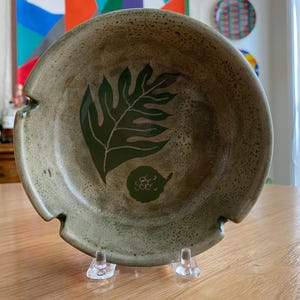 May include: A handmade, speckled ceramic bowl with a leaf design. The bowl is a muted green color with a dark green leaf and fruit design. The bowl has three cut-out sections on the rim and is displayed on a clear stand.