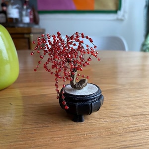 May include: A miniature bonsai tree sculpture with a wire trunk and branches adorned with red beads. The tree sits in a small, black, decorative pot. The bonsai is a unique decorative item.