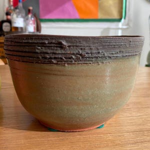 May include: A handmade ceramic bowl with a textured, dark brown rim and a light brown and green body. The bowl is round and sits on a wooden surface. The bowl is a decorative item.
