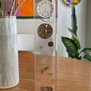 May include: A clear resin sculpture featuring vintage watch parts. The rectangular piece has a red base and an arched top. Inside, a clock face and various gears and springs are suspended, creating a unique decorative item.