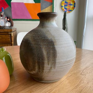 May include: A handcrafted ceramic vase with a rounded body and a narrow neck. The vase features a neutral color palette with shades of beige, brown, and cream. The design includes vertical brown streaks, adding visual interest to the piece.