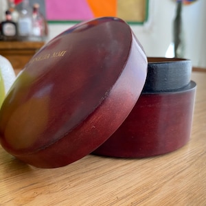 May include: A partially open, oval-shaped, wooden box with a dark red finish. The lid is slightly ajar, revealing the interior and a black rim. The box sits on a wooden surface. The text "ANGELA MAIT" is visible on the lid.