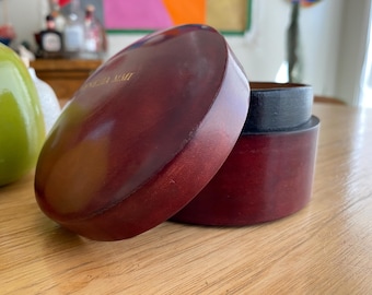 May include: A partially open, oval-shaped, wooden box with a dark red finish. The lid is slightly ajar, revealing the interior and a black rim. The box sits on a wooden surface. The text "ANGELA MAIT" is visible on the lid.