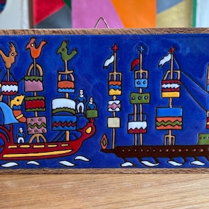 May include: A colorful ceramic tile with a folk art design. The artwork features two stylized ships with various shapes and figures in red, blue, orange, green, and yellow. The tile is framed in a wooden border.