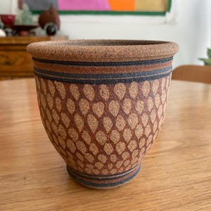 May include: A textured ceramic planter with a tapered base and a wide rim. The exterior features a repeating pattern of light brown teardrop shapes against a darker brown background, accented by two horizontal black bands near the top and bottom.