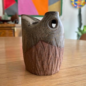 May include: A ceramic vessel featuring a dual-tone finish with a dark, speckled brown glaze on the upper portion and a textured, reddish-brown clay base. The object has a distinct asymmetrical rim and a circular cutout near the top edge.