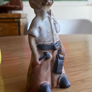 May include: A ceramic figurine of a man in work clothes, with a white shirt, brown pants, and a black belt. The figurine has a mustache and is holding a hammer and a tool bag. The figure is standing on a wooden surface.