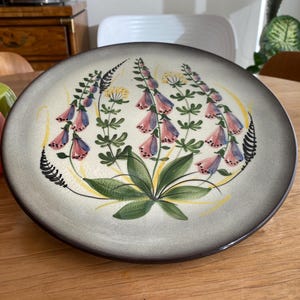 May include: A round ceramic plate with a floral design. The plate features a light gray background with a dark brown rim. The floral design includes pink, blue, and yellow flowers with green leaves. The plate is on a wooden table.