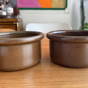 May include: Two brown ceramic ramekins with a slightly textured surface. The ramekins are oval-shaped with a rounded rim. They are sitting on a wooden table. The ramekins are likely used for baking or serving individual portions of food.