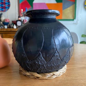 May include: A black, handcrafted ceramic vase with a textured leaf design around the base. The vase sits on a round, woven straw coaster. The vase has a rounded body and a slightly flared rim. Decorative home decor.