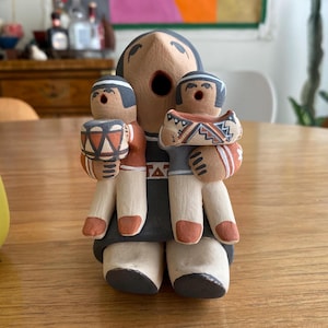 May include: A handcrafted ceramic figurine depicting a central figure flanked by two smaller figures. The central figure has an open mouth and is wearing a dark gray robe. The smaller figures hold drums and a decorated object. The color palette includes tan, orange, black, and white.